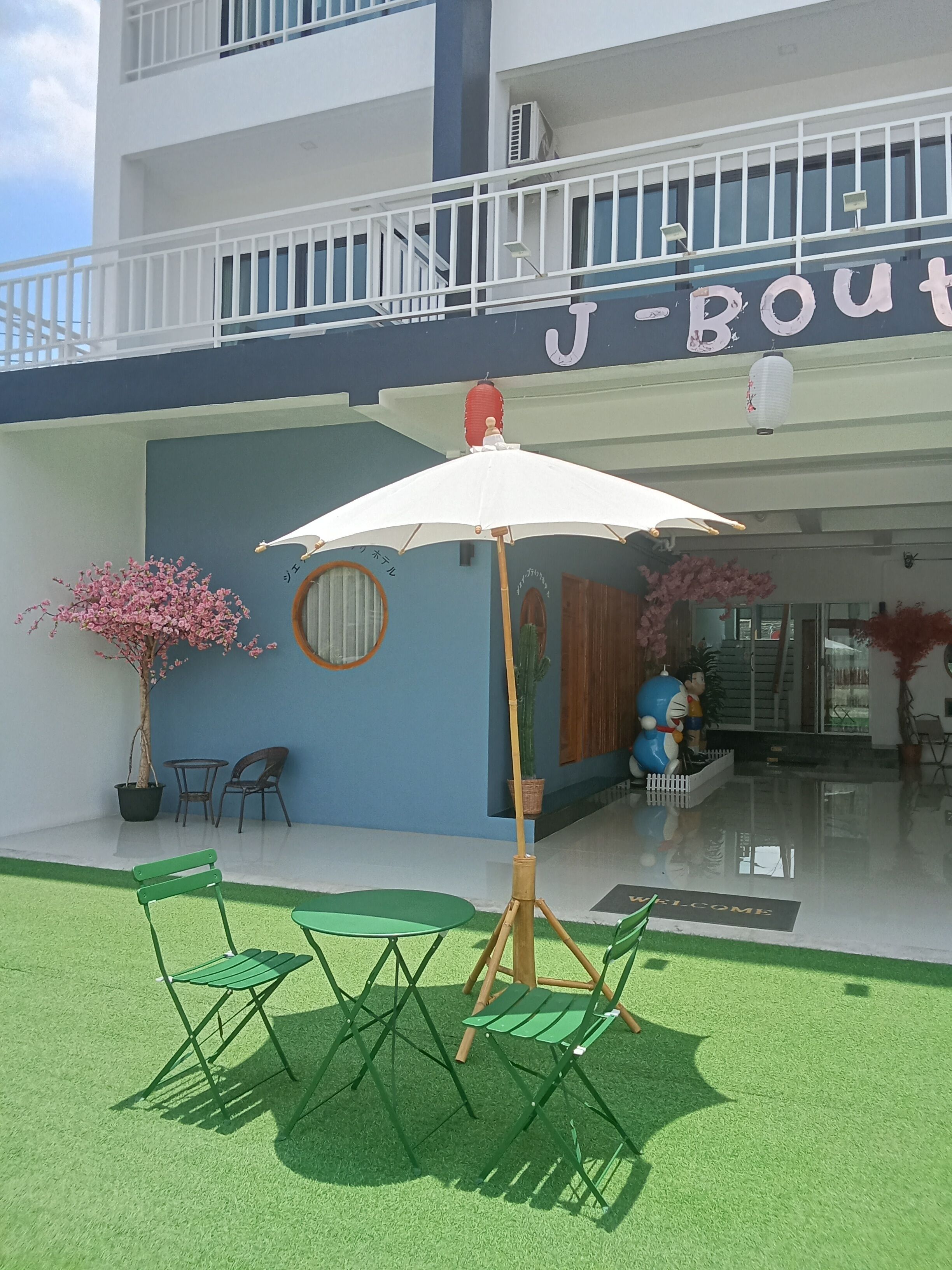 Entrada de hotel con césped verde, una sombrilla blanca y un letrero que dice "J-Boutique".