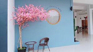 A room with a blue wall, a round mirror, a potted tree with pink flowers, and two wicker chairs. - J-Boutique Hotel (Khon Kaen)