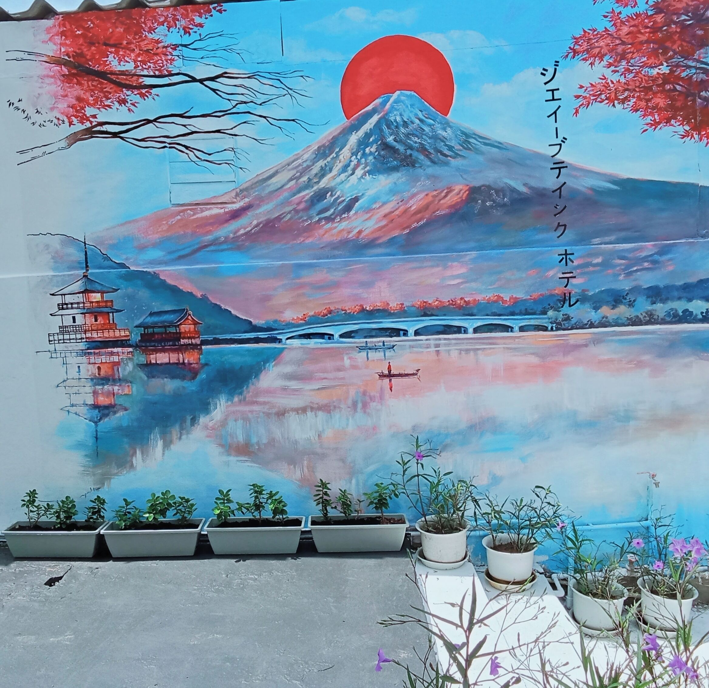 Une fresque représentant le mont Fuji, avec un pont et une pagode, le tout entouré de plantes en pot.