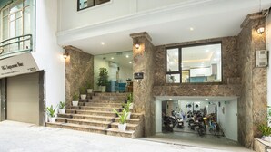 A hotel entrance with a stone facade, a staircase leading to the upper floor, and a visible parking area with motorcycles.