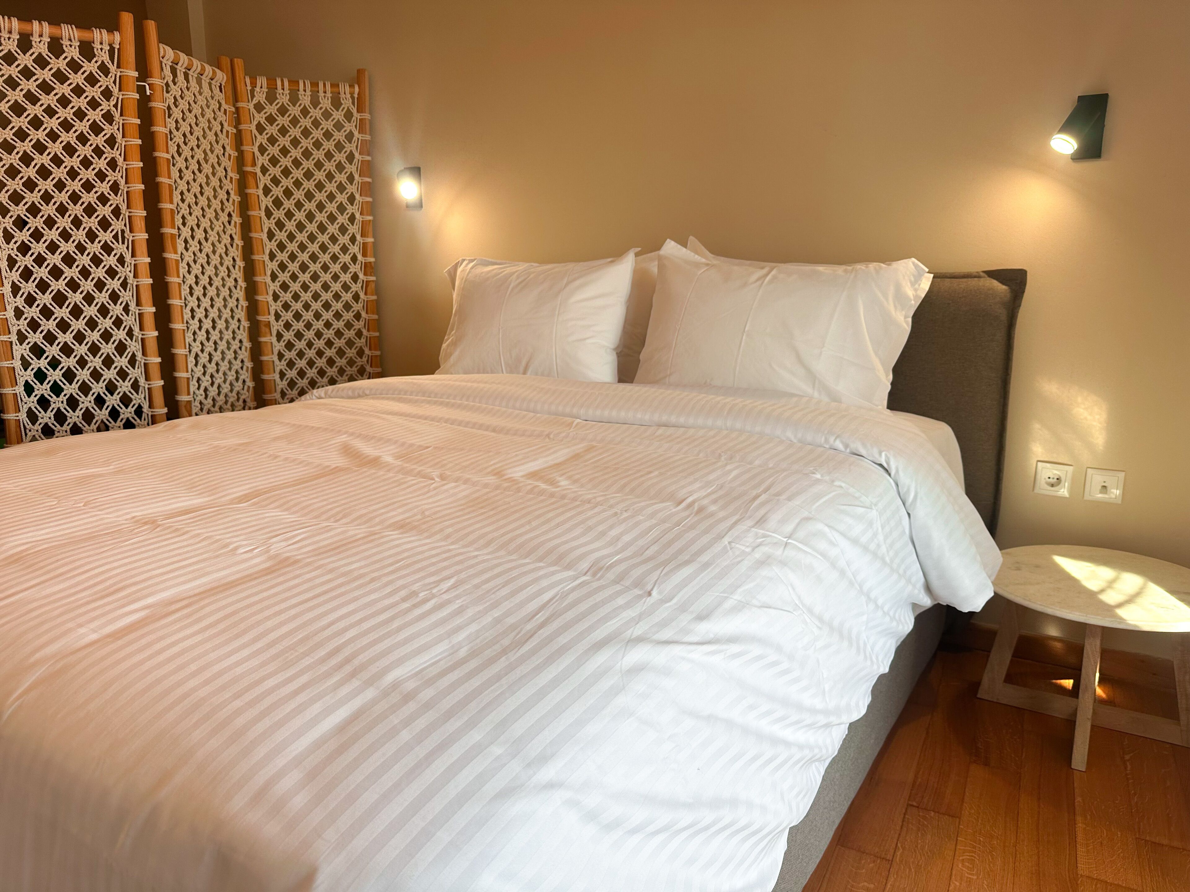A neatly made bed with white linens, a small round table beside it, and a wall-mounted light fixture.