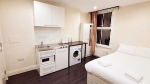 Een kleine hotelkamer met een bed, een kitchenette, een wasmachine en een raam met gordijnen.