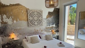 Quarto com uma cama grande, roupa de cama branca, almofadas decorativas, luminária fixada na parede e vista para um jardim de cactos através de uma porta de vidro.