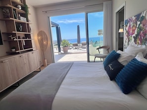 Un dormitorio con cama, un aparador de madera, una planta en maceta y vista al mar a través de una puerta corredera.
