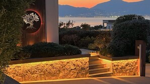 Um edifício moderno com fachada de pedra, escadaria de madeira e vista para a água e montanhas ao pôr do sol.