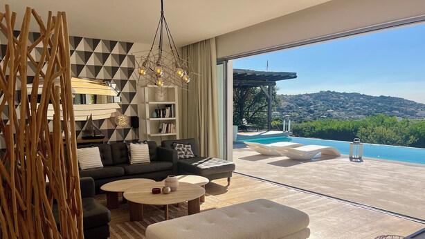 Sala de estar moderna com sofá, mesa de centro, repouso para os pés, estante e vista para uma piscina e colinas.