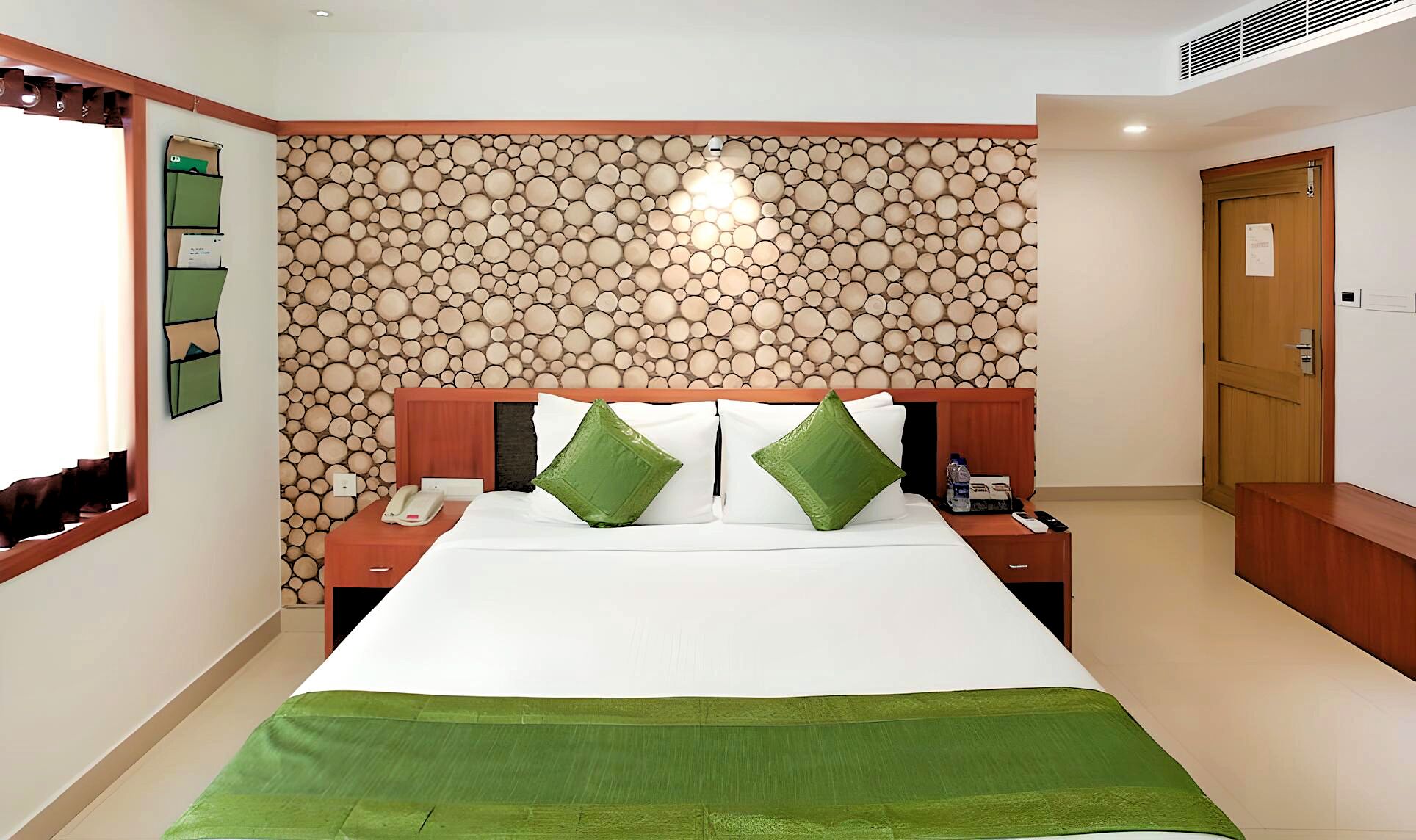 A hotel room with a bed, green pillows, a nightstand with a phone, and a wall with a circular pattern.