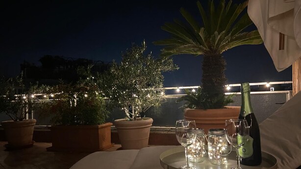 Una terraza en la azotea con plantas en macetas, una mesa con copas de vino y una botella, y luces colgantes.