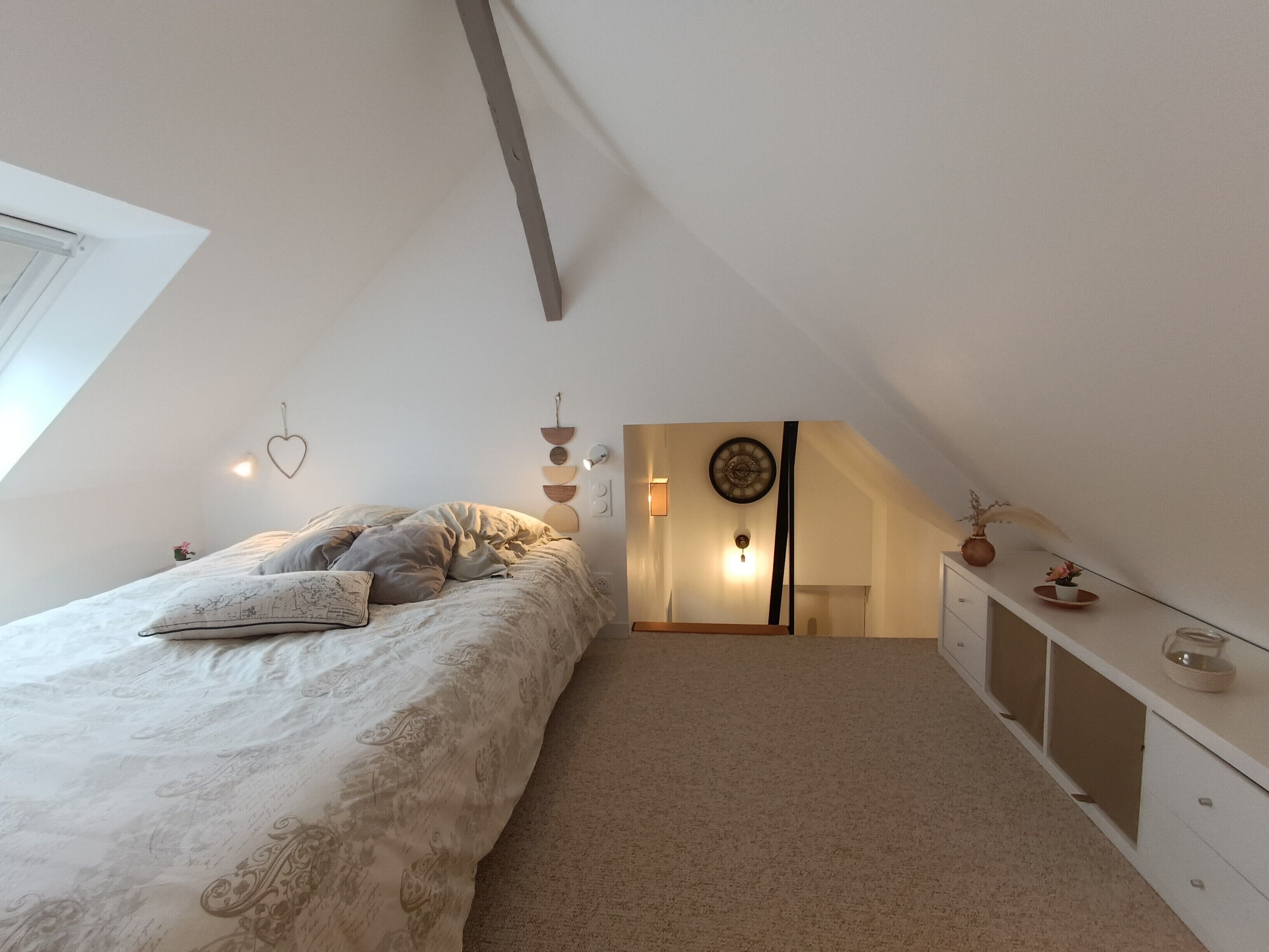 A bedroom with a sloped ceiling, a bed with patterned bedding, a bedside table with decorative items, and a wall-mounted light fixture.