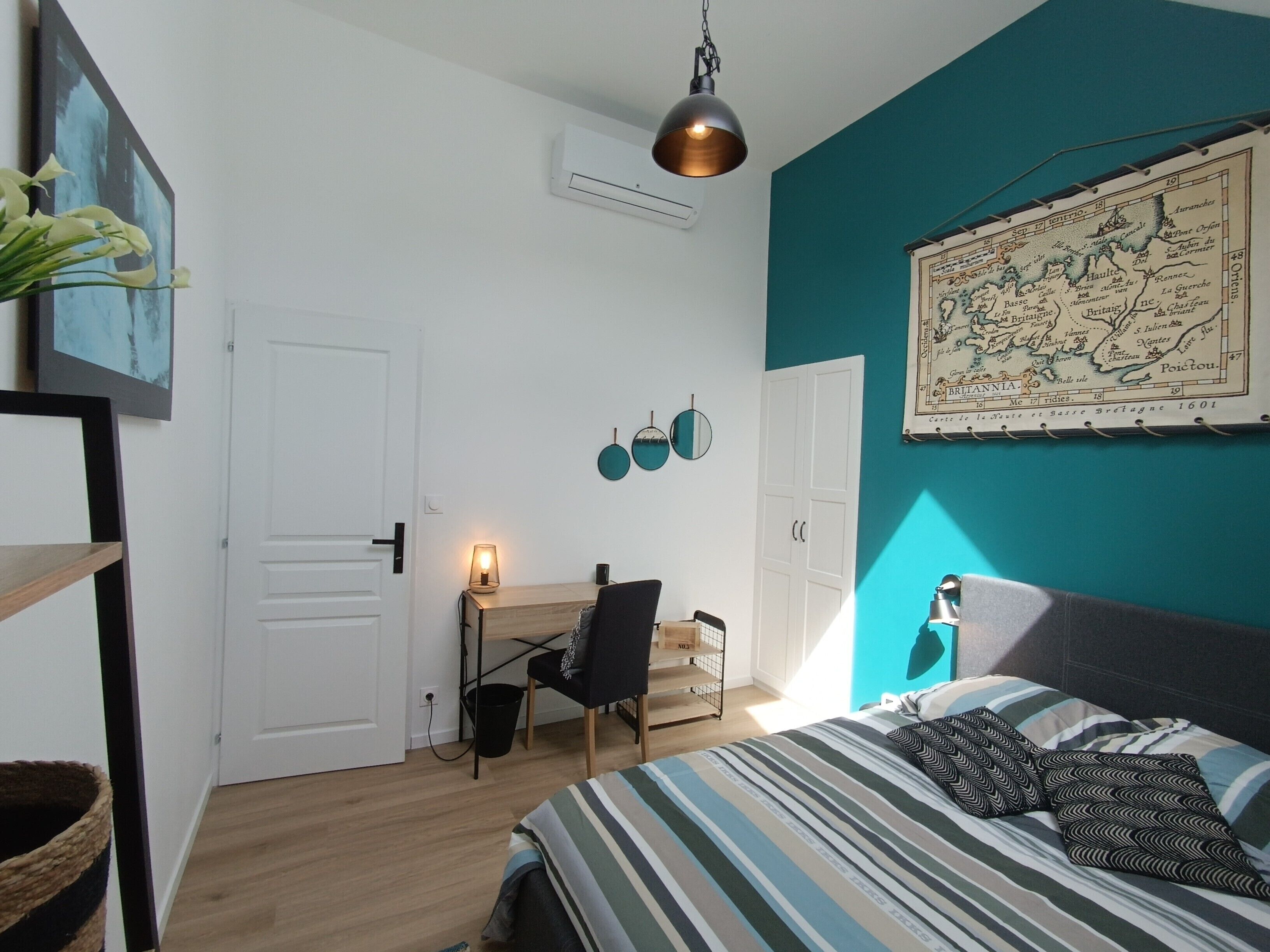 A bedroom with a bed, a desk, and a chair. There is a map on the wall and a hanging light fixture.
