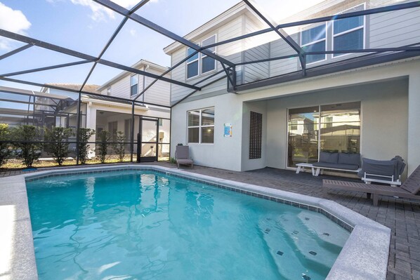 Ein Schwimmbad mit Glasveranda, eine Terrasse mit Sitzgelegenheiten und ein Blick auf ein Haus im Hintergrund.