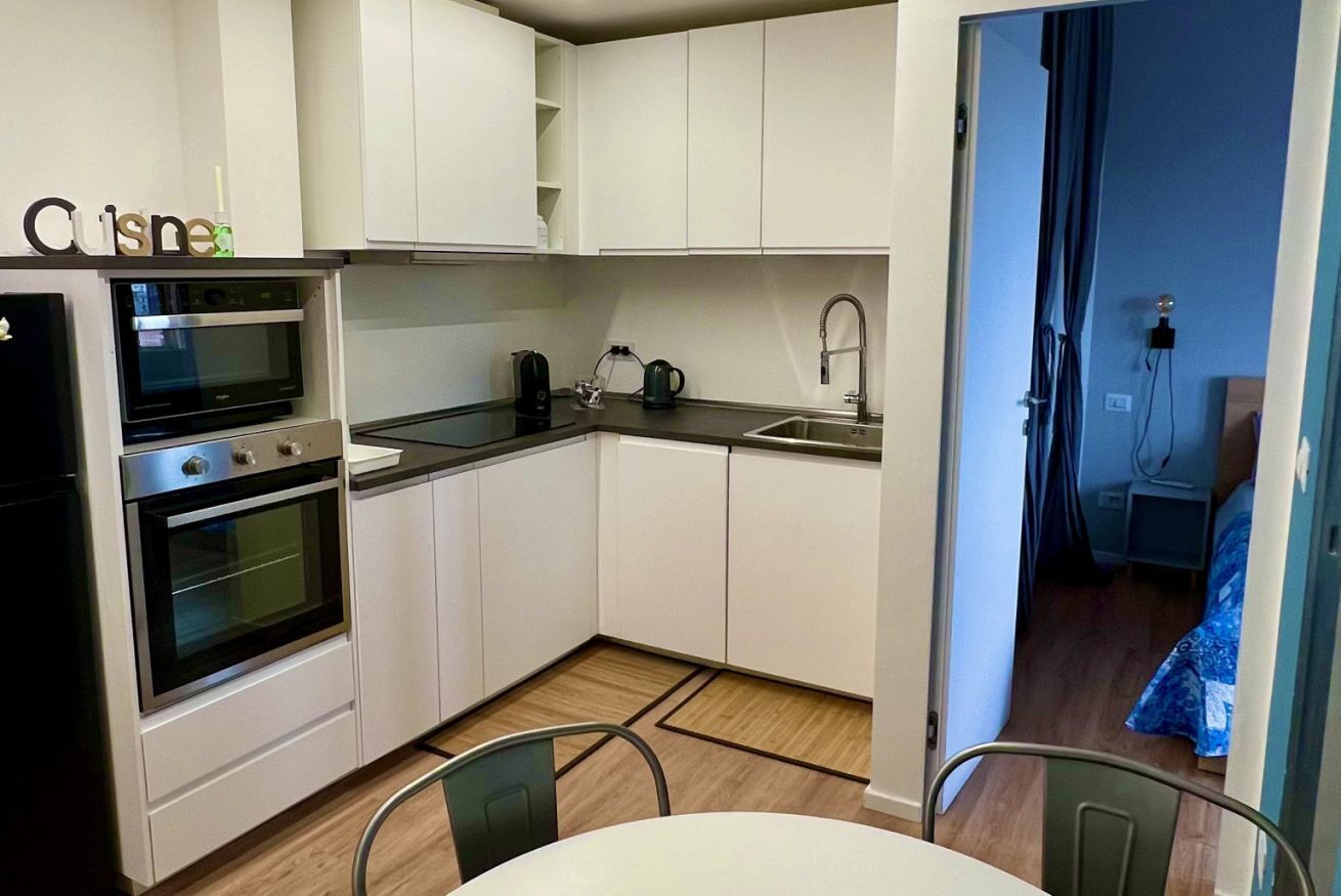 Cocina compacta con armarios blancos, horno empotrado, pileta y comedor con mesa y sillas.