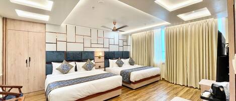 A modern hotel room with two beds, a wooden floor, a desk with a remote control, and a large window with curtains.
