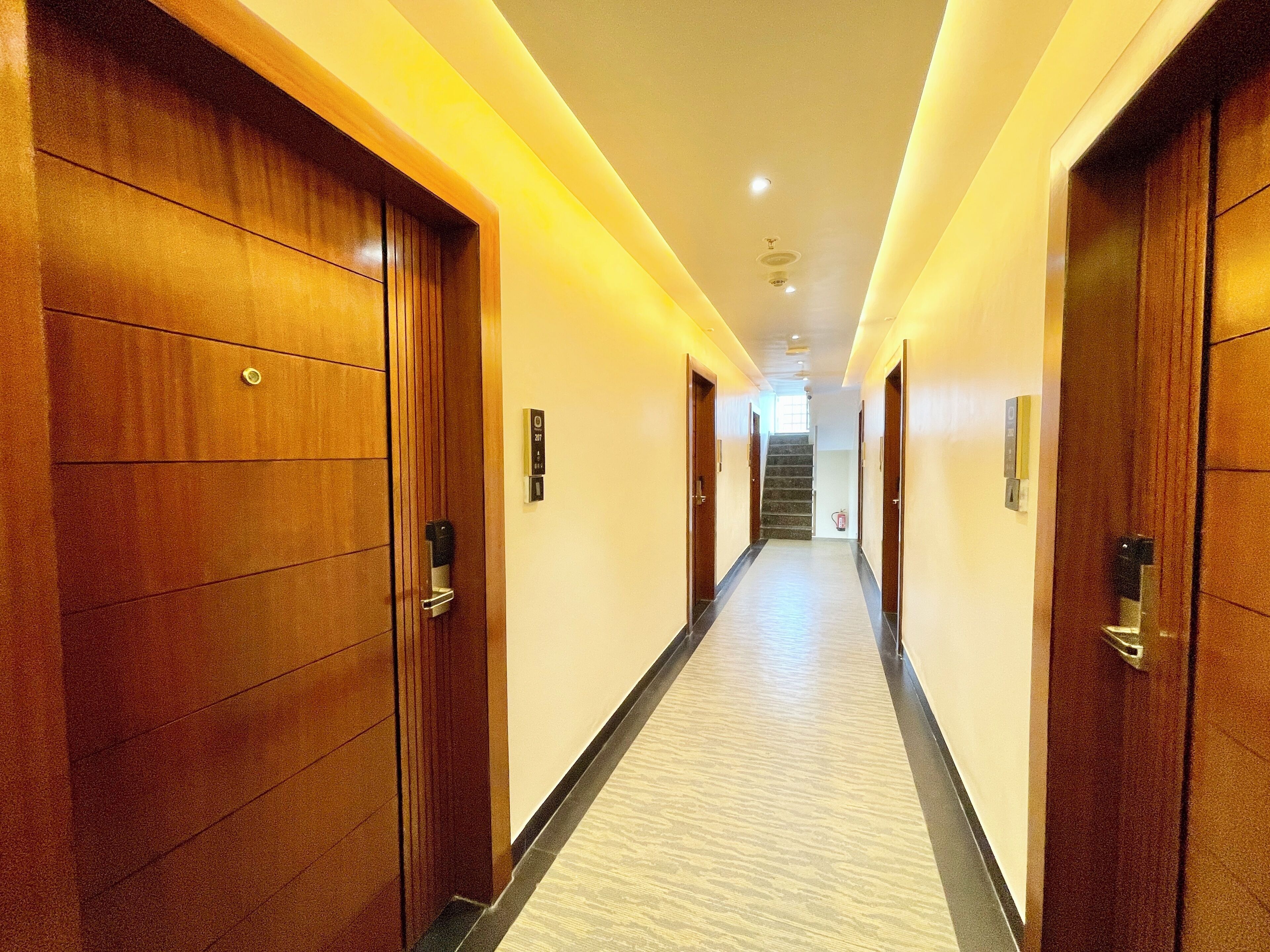 A hotel hallway with wooden doors, a carpeted floor, and a staircase at the end.