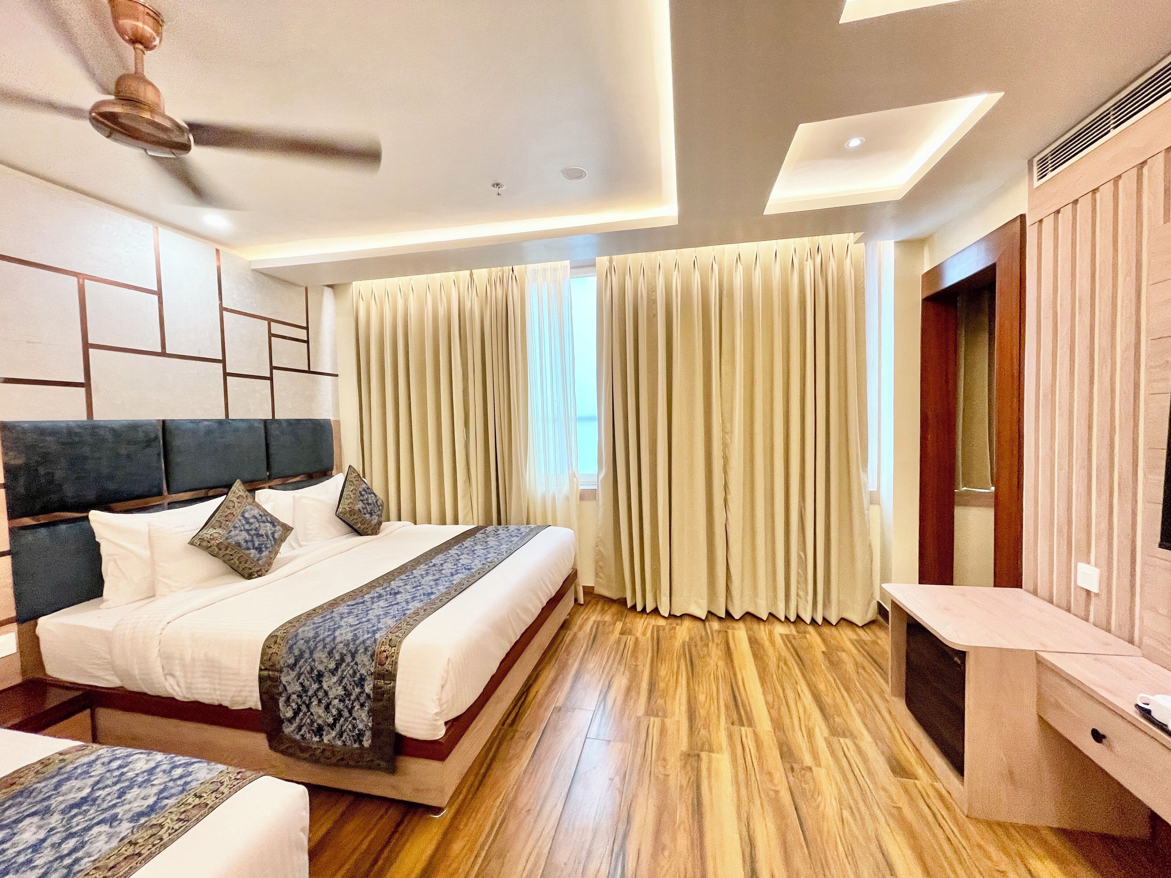 A modern hotel room with a large bed, wooden flooring, a ceiling fan, and a built-in TV cabinet.