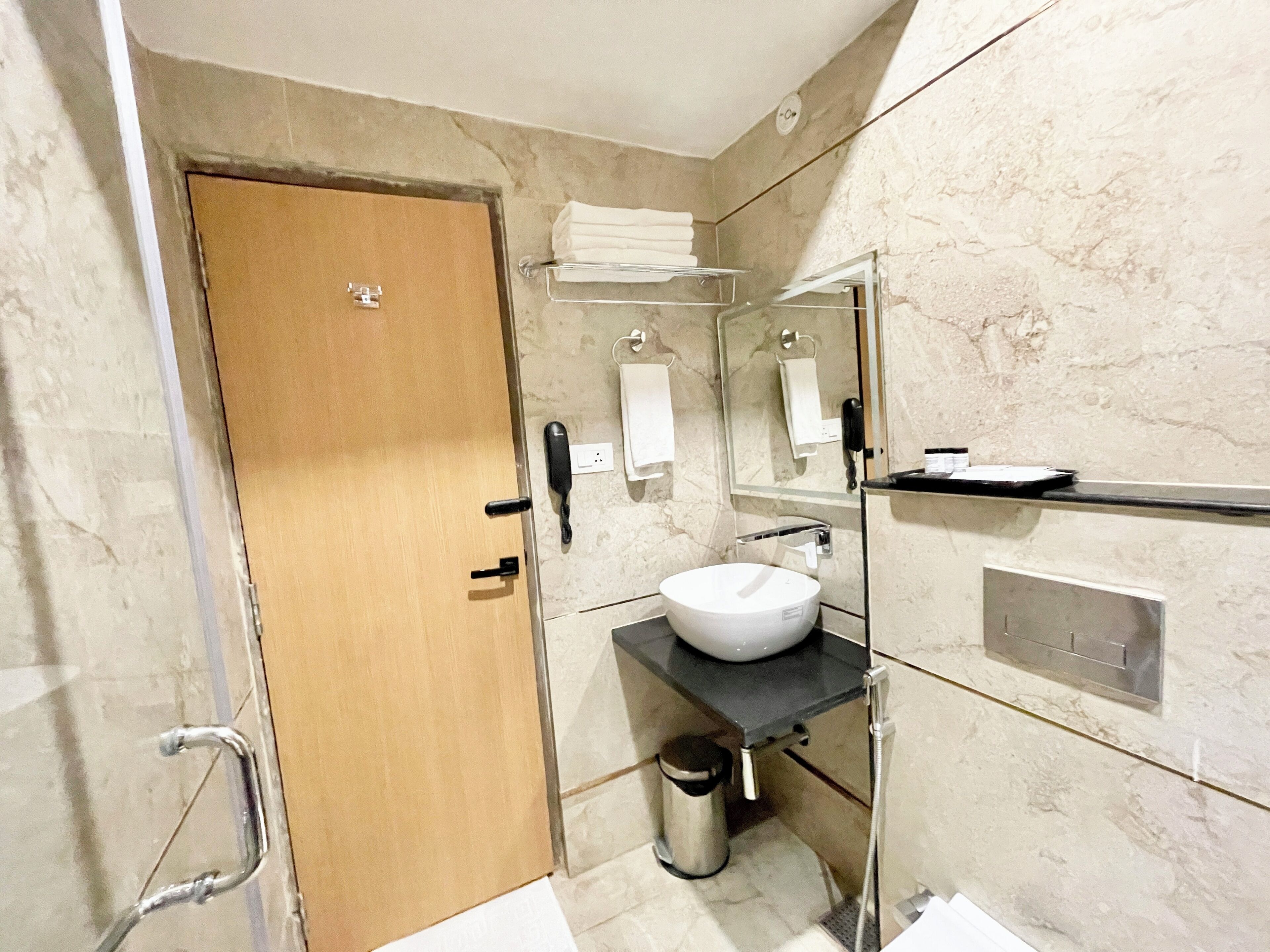 A modern bathroom with a wooden door, a white sink, a glass shower enclosure, a towel rack, and a trash bin.
