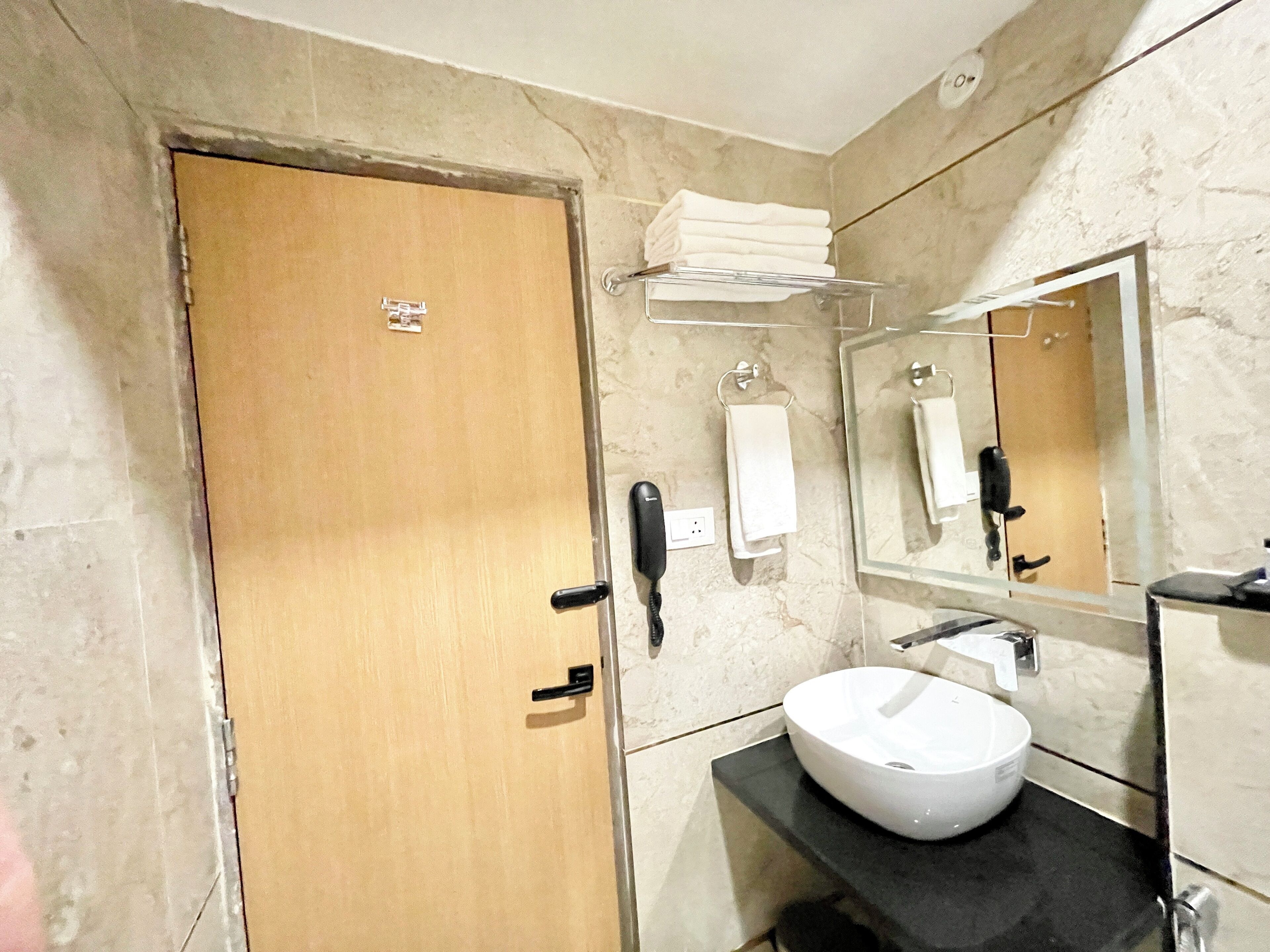 Un baño con puerta de madera, lavamanos blanco, espejo, toallero y teléfono.
