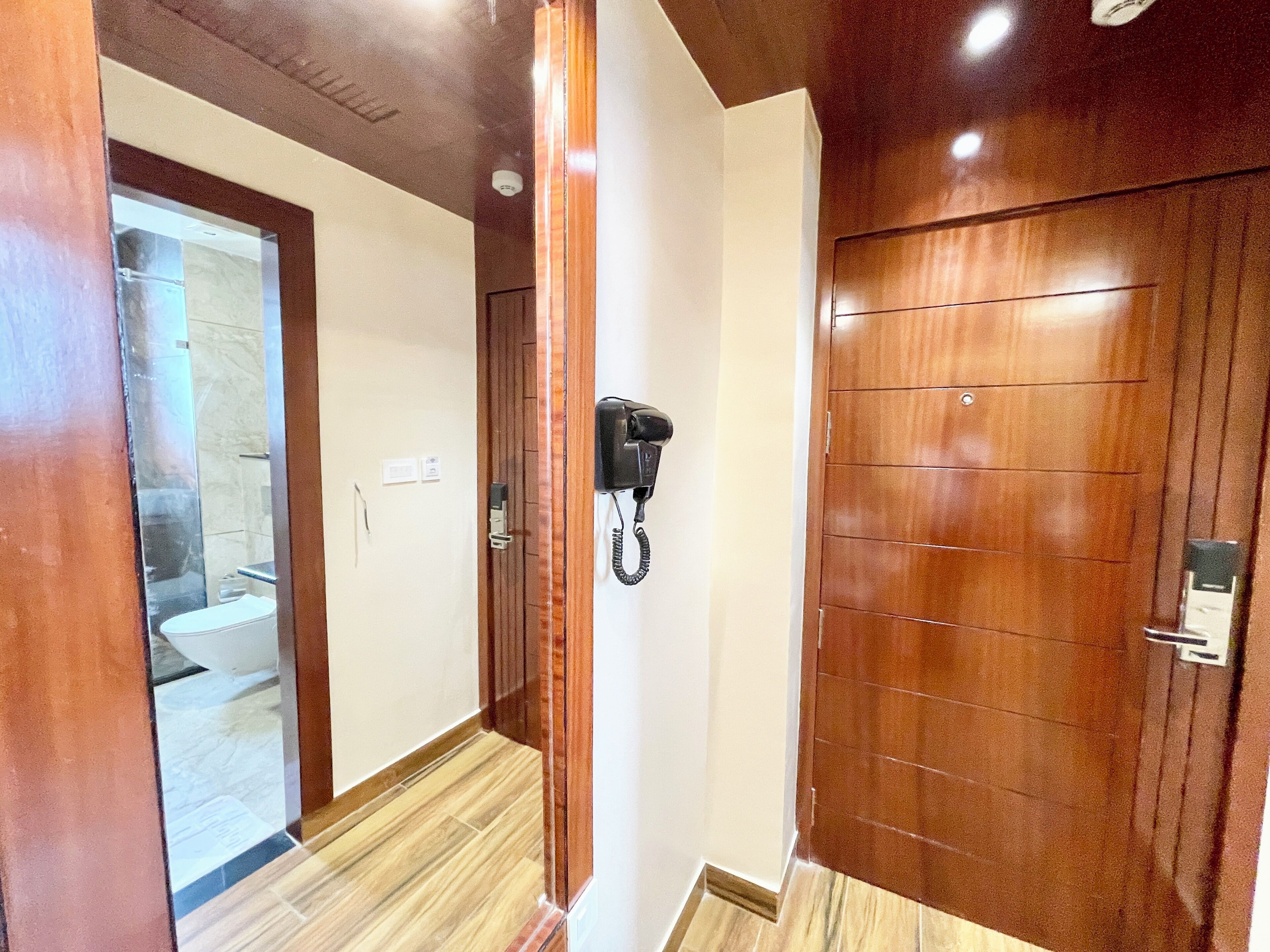 Un pasillo con una puerta de madera, un teléfono y un espejo que refleja un baño.