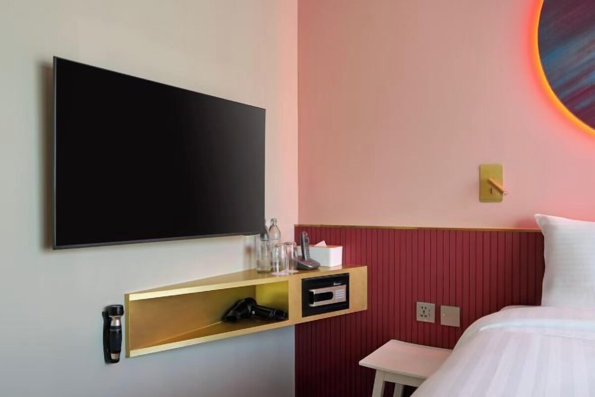 A hotel room with a flat-screen TV mounted on the wall, a bedside table with a lamp, and a bed with white bedding.