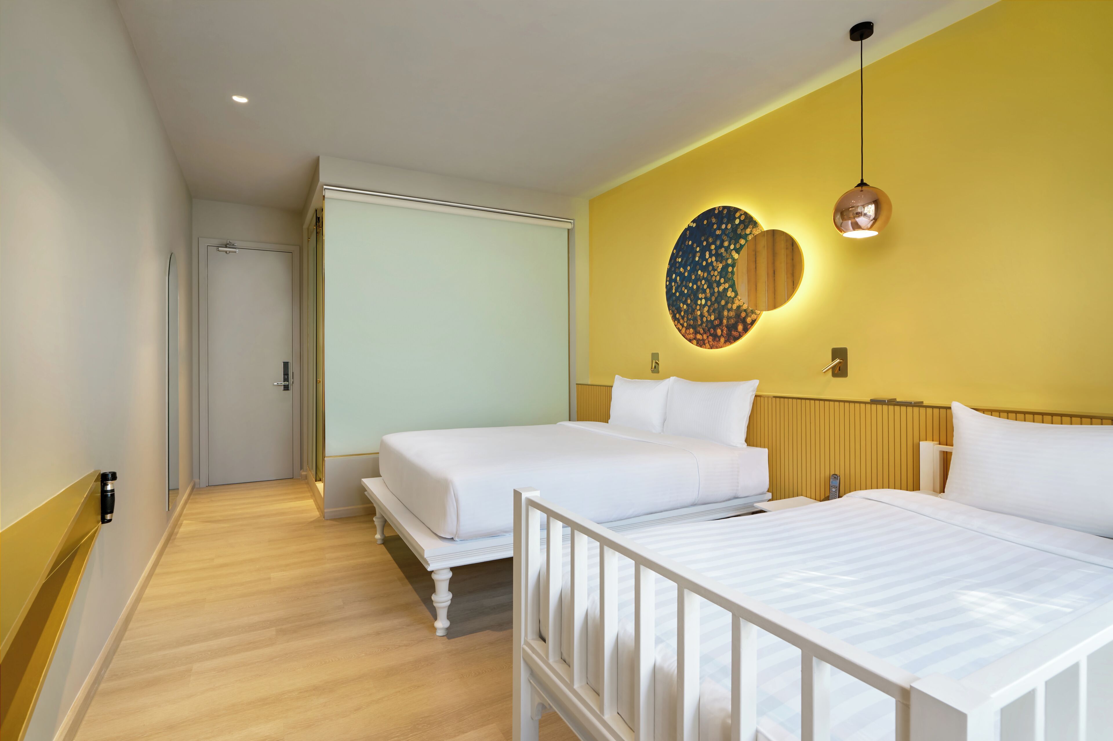 A hotel room with two beds, a wooden floor, a yellow accent wall, and a circular wall decoration.