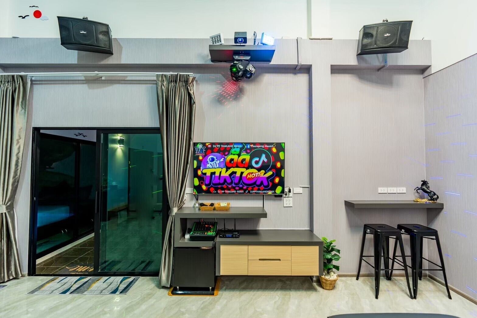 Un salon moderne doté d’un téléviseur à écran plat diffusant un remix de musique thaïlandaise, d’une porte d’entrée en verre, d’un petit meuble en bois et de deux tabourets de bar noirs.