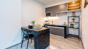 A modern kitchen with dark cabinets, a black countertop, and a built-in microwave. There is a small island with a vase and a plant, and a shelf with kitchen items. - 58-20 Pool Villa (Cha-am)