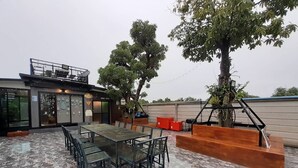 A modern outdoor patio with a glass table and chairs, surrounded by potted plants and a large tree. - Intouch Poolvilla (Cha-am)