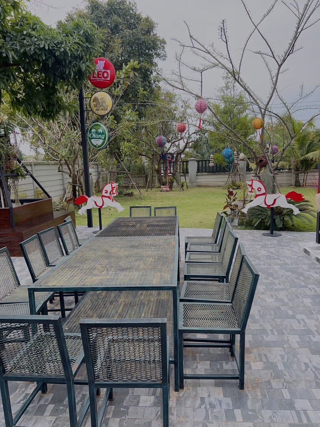 Un área de estar al aire libre con mesas y sillas de metal, rodeada de árboles y elementos decorativos.