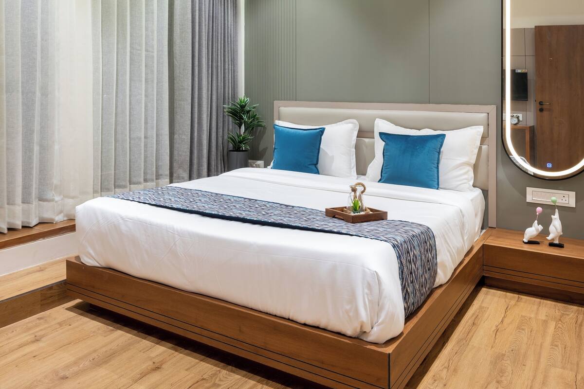A modern bedroom with a large bed, wooden flooring, a bedside table with a mirror, and a window with curtains.
