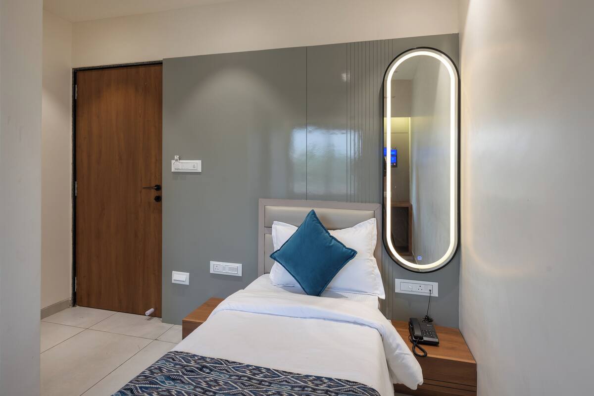 A hotel room with a bed, a bedside table with a phone, a mirror with lights, a door, and a wall socket.
