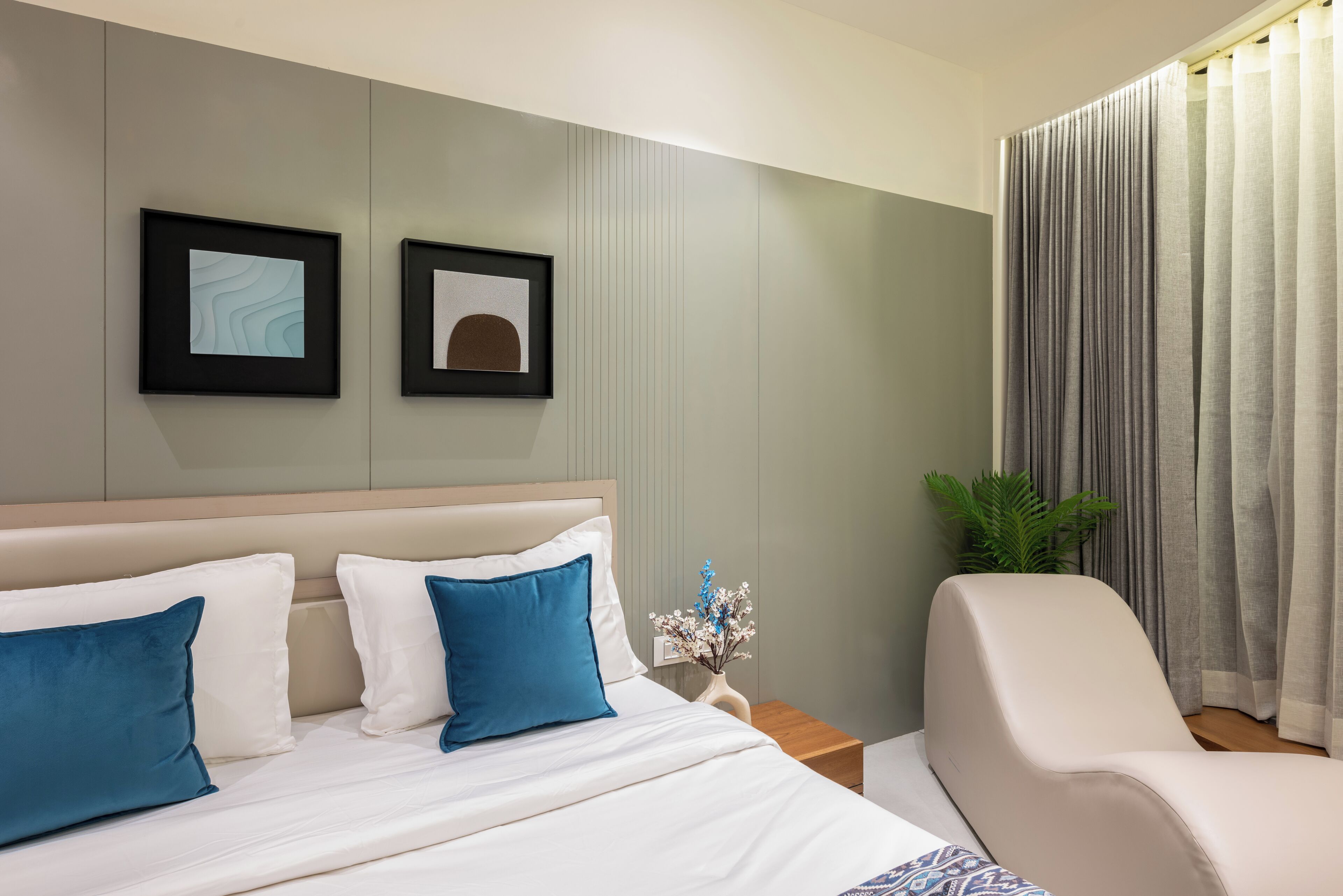 A modern hotel room with a bed, a nightstand, a chair, and two framed pictures on the wall.