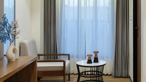 A modern living room with a wooden floor, a white sofa, a round black table with two decorative items, a vase with blue flowers, and a large window with grey curtains. - The Zuree Hotel (Gandhinagar)