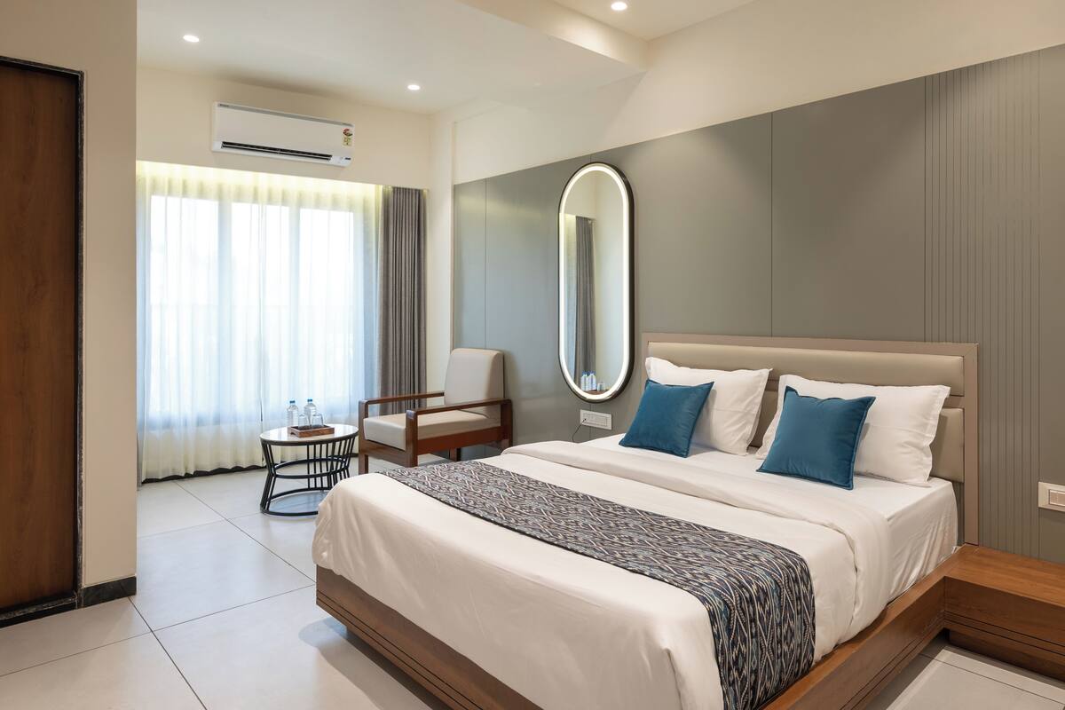 A modern hotel room with a large bed, a round mirror, a small table with two bottles, and a chair.