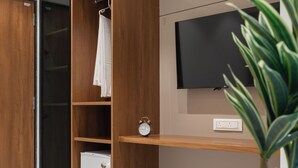 A modern bedroom with a wooden wardrobe, a flat-screen TV mounted on the wall, a small shelf, and a potted plant. - The Zuree Hotel (Gandhinagar)