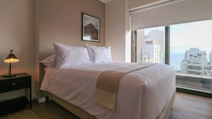 A neatly made bed with white linens and a throw blanket, a bedside lamp, a framed picture on the wall, and a large window with a view of buildings and the sea. - w* | Exlusive 3BR w/ Balcony Near Larcomar (Miraflores)