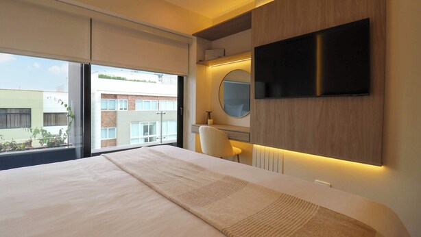 A modern hotel room with a large bed, a flat-screen TV mounted on the wall, a round mirror, and a view of the outside. - w* | Elegant 2BR w/ Balcony Near Larcomar (Miraflores)