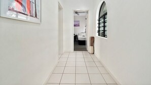 Un pasillo con piso de baldosas blancas, una obra enmarcada a la izquierda y una ventana con celosía metálica a la derecha.