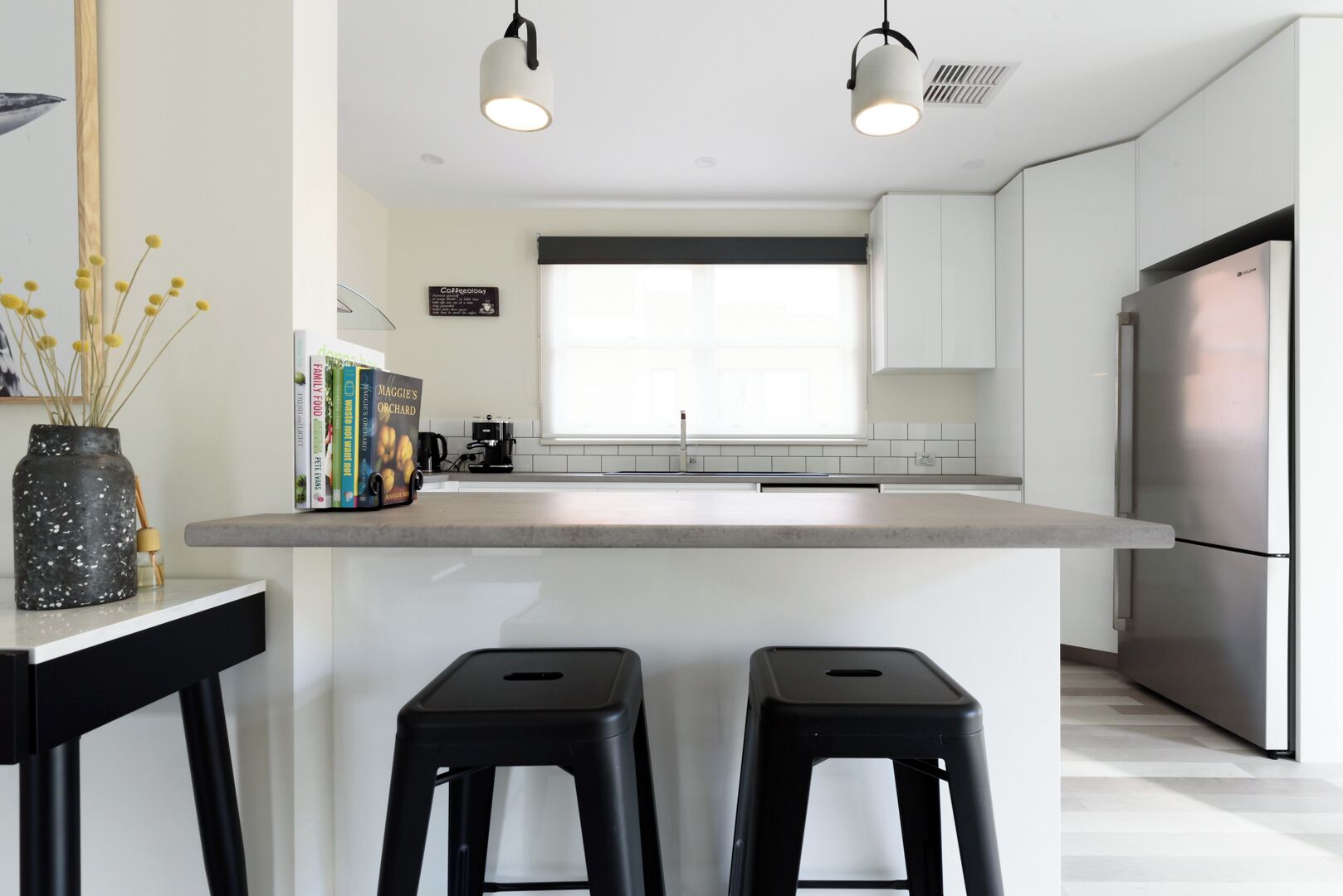 A modern kitchen with a central island, two black stools, a coffee machine, and a refrigerator.