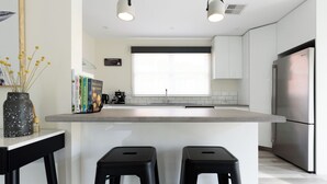 Una cocina moderna con una isla central, dos taburetes negros, una cafetera y un refrigerador.