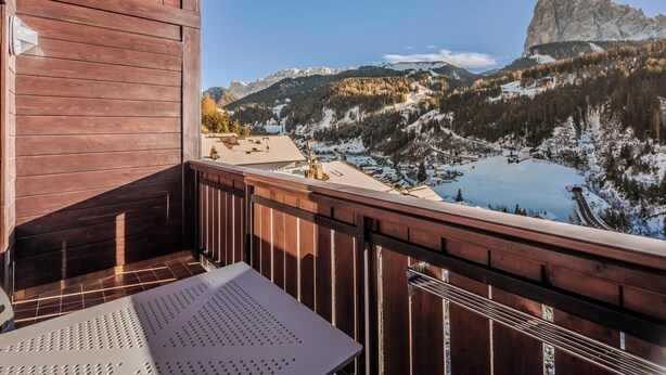Un balcón de madera con mesa y sillas, con vista a un paisaje montañoso que incluye un lago y una destacada formación rocosa.