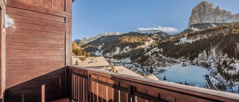 A wooden balcony with a table and chairs, overlooking a mountainous landscape with a lake and a prominent rock formation.