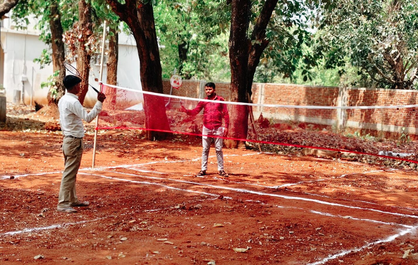 A person playing badminton on a dirt court with trees in the background.