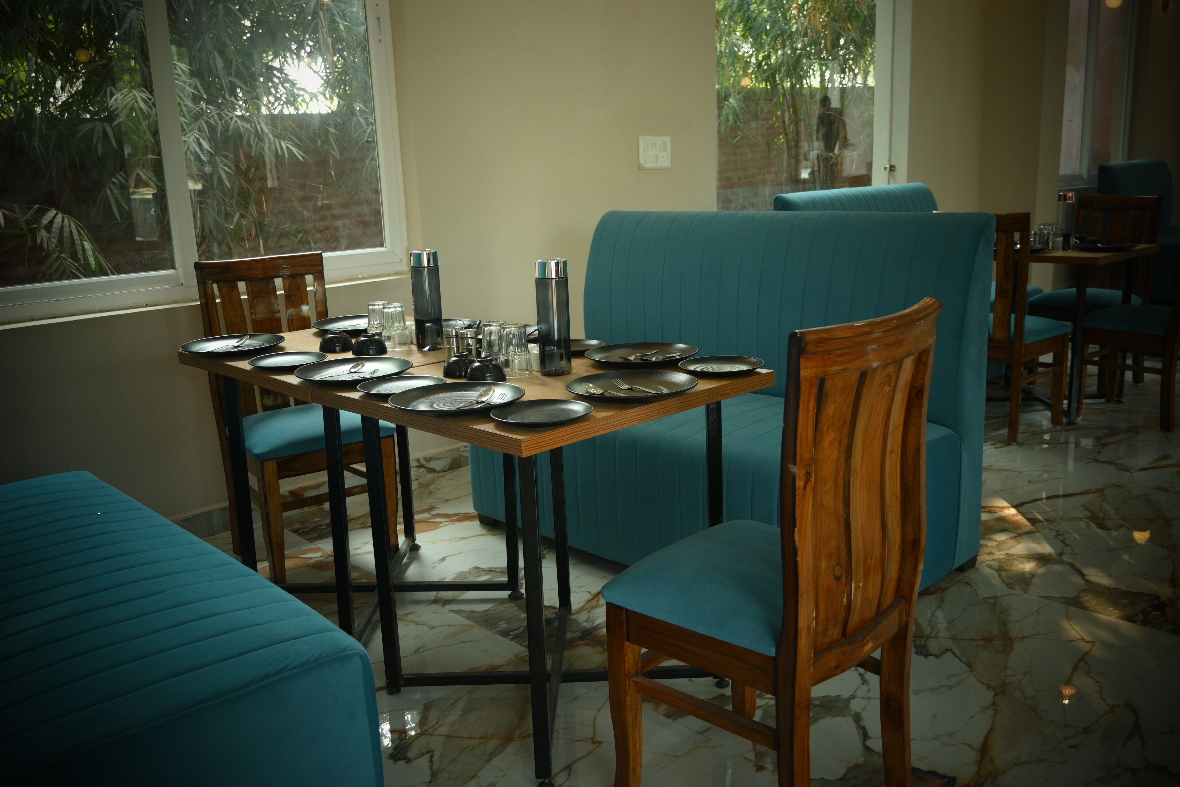 A dining area with a table set for four, teal upholstered chairs, and a view of greenery through the windows.