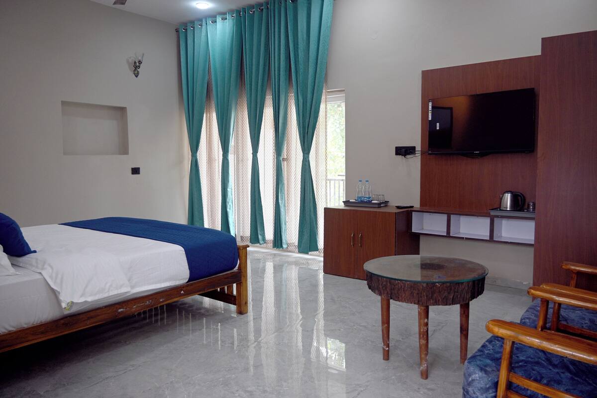 Quarto de hotel com cama, mesa redonda pequena, televisão presa a um painel de madeira e armário com chaleira.