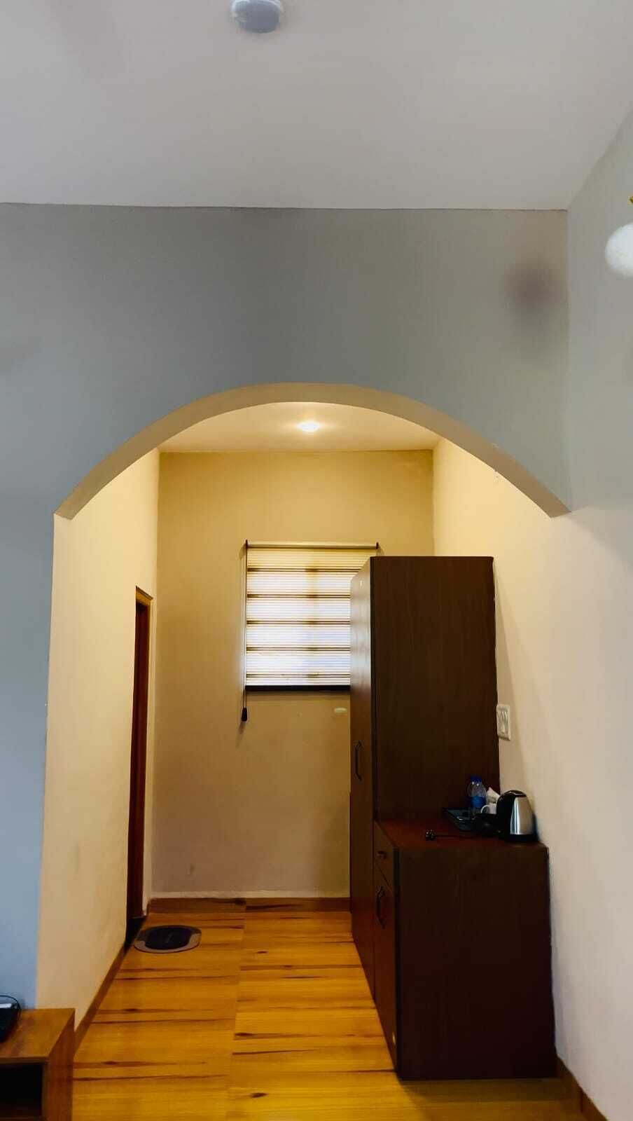 A narrow hallway with wooden flooring, a window with blinds, and a small cabinet with a kettle on top.