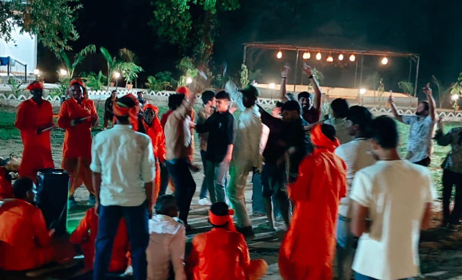 A group of people gathered outdoors at night, some wearing red, under a canopy with lights.