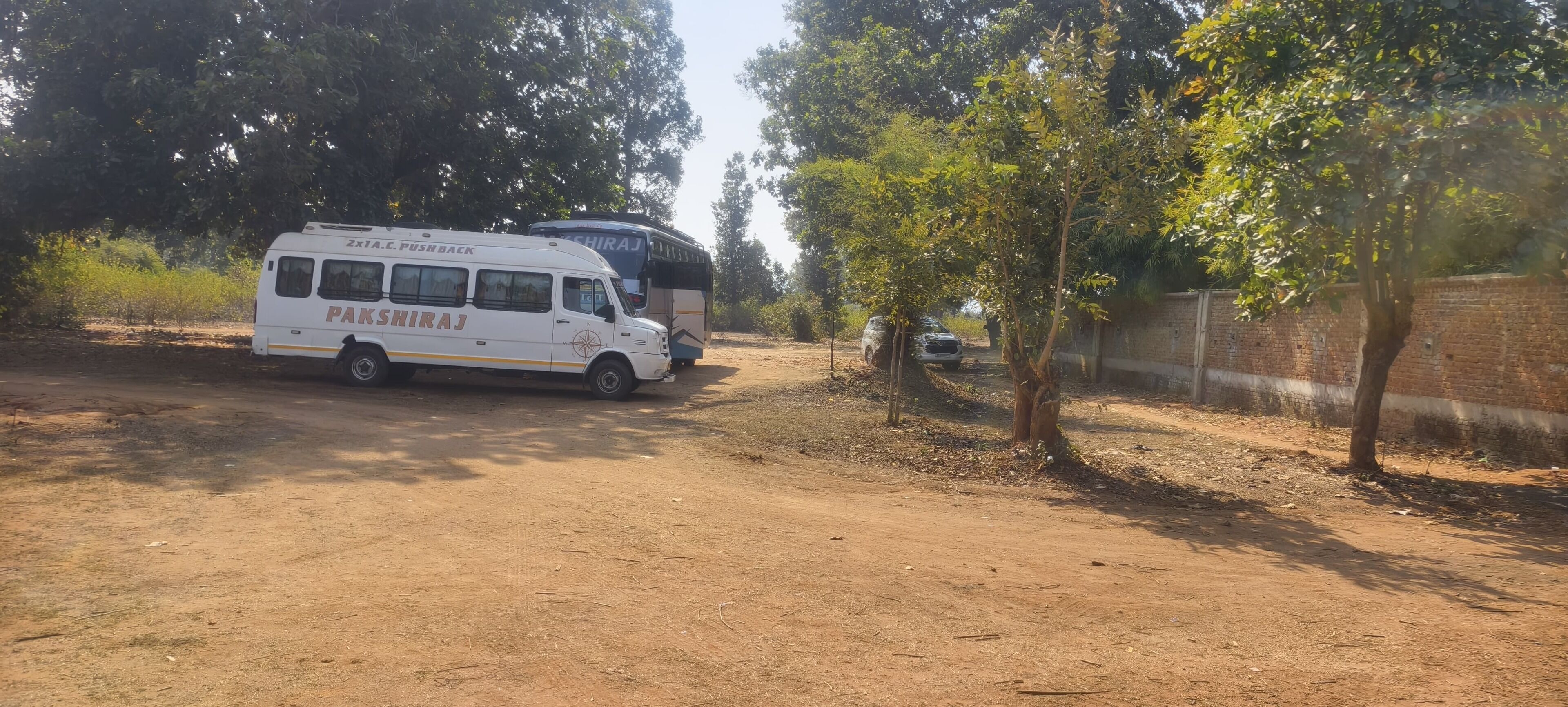 A white van labeled "PAKSHIRAJ" and a bus parked on a dirt road with trees and a brick wall in the background.