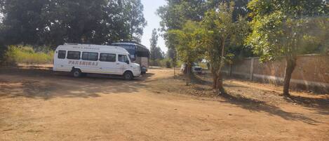 A white van labeled "PAKSHIRAJ" and a bus parked on a dirt road with trees and a brick wall in the background.