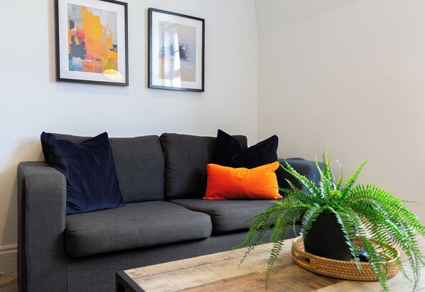 En grå sofa med sorte og orange puder, et indrammet abstrakt maleri og en grøn bregne på et træbord.