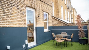 A brick building with a dark blue exterior, a glass door, and a small patio with artificial grass and wicker chairs. - Brixton/2bed Flat/privateterrace/tv/wifi (London)
